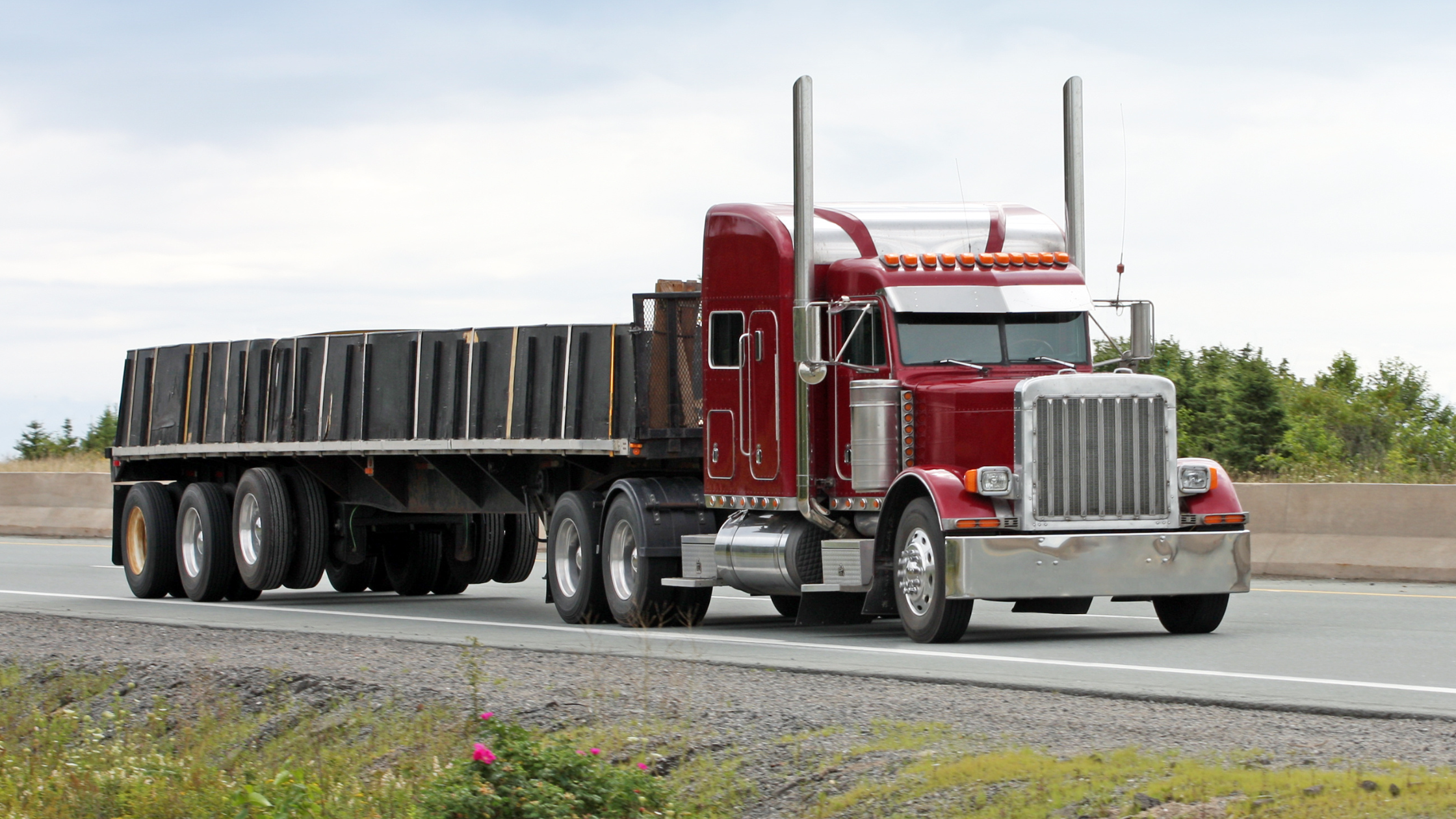 a maroon semi-truck driving on the highway with an open deck trailer.